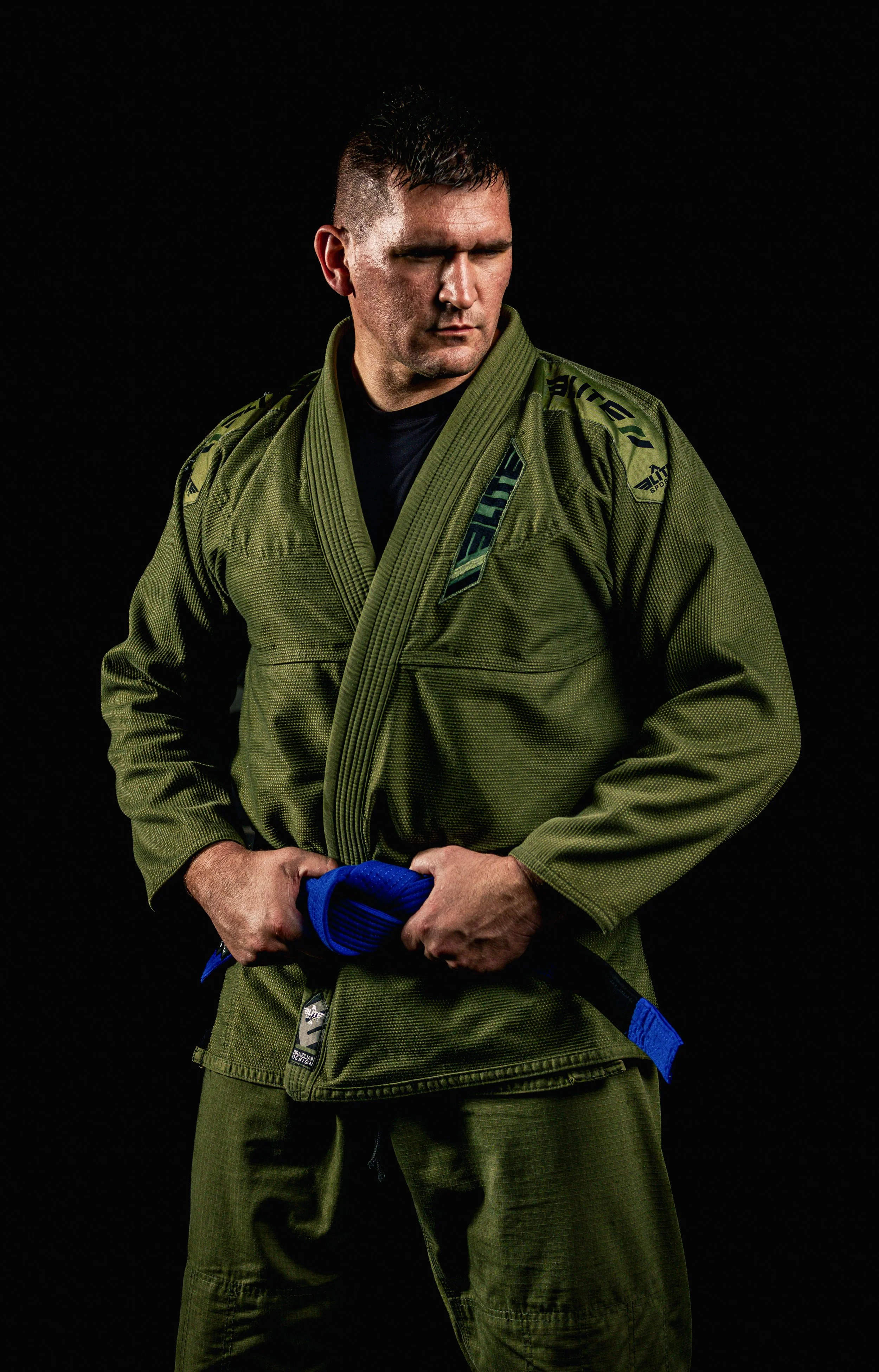 Athlete portrait of a martial artist in a green gi holding a blue belt against a black background.