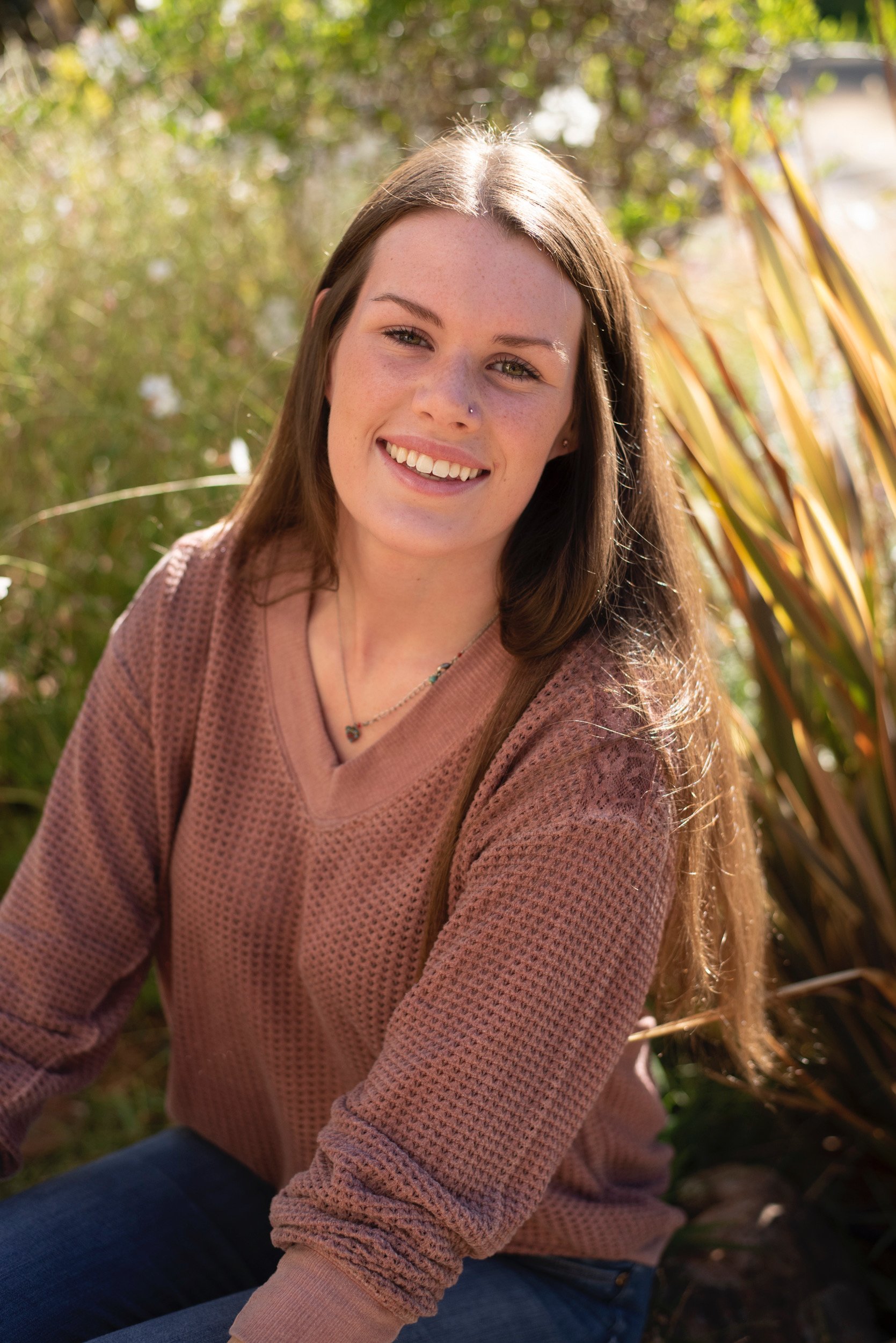 Young woman with long brown hair, wearing a pinkish-brown sweater and blue jeans, sitting outdoors in a natural setting with green foliage and sunlight.