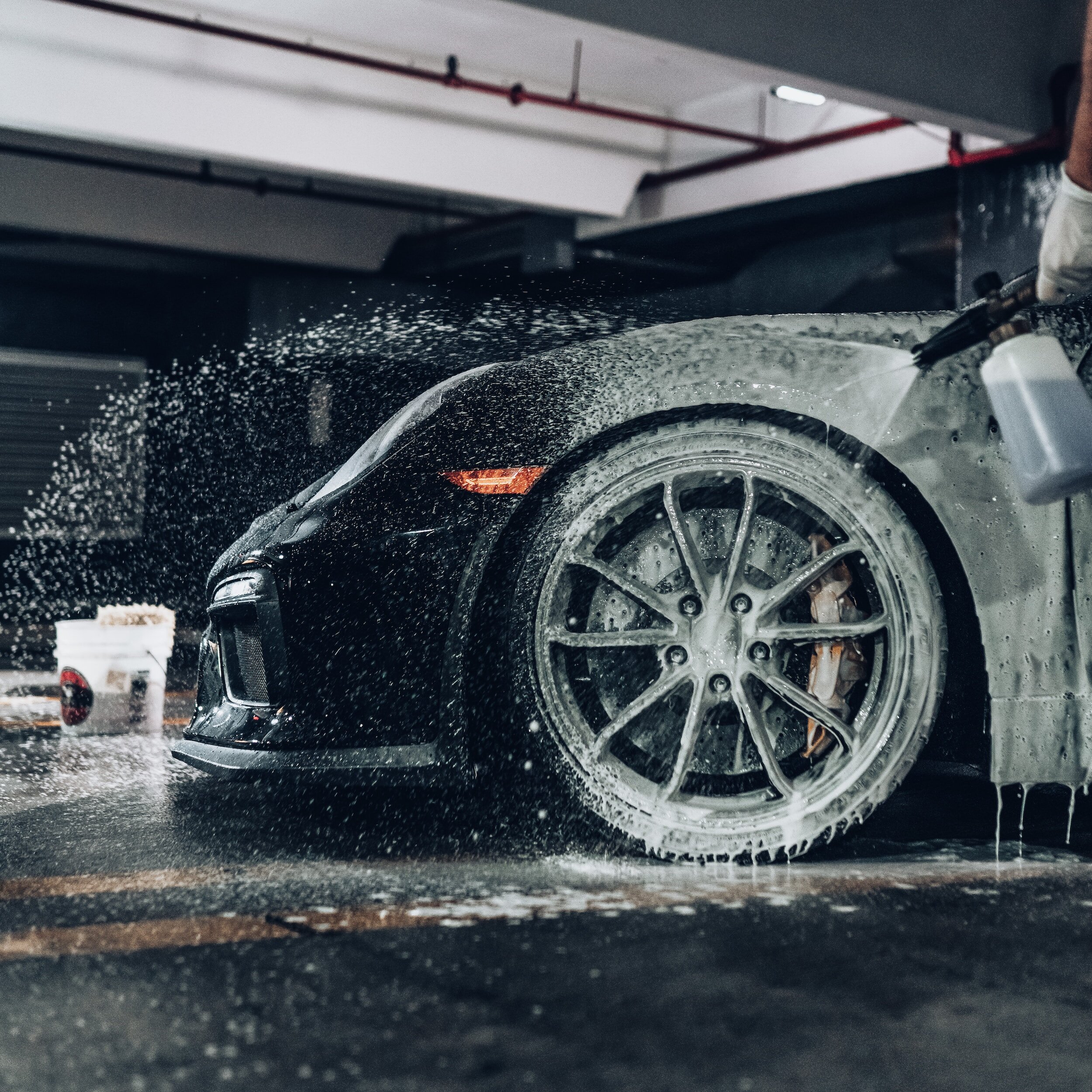 A close-up view of a black car being washed, with water and soap visible on the car's surface and silver alloy wheels.