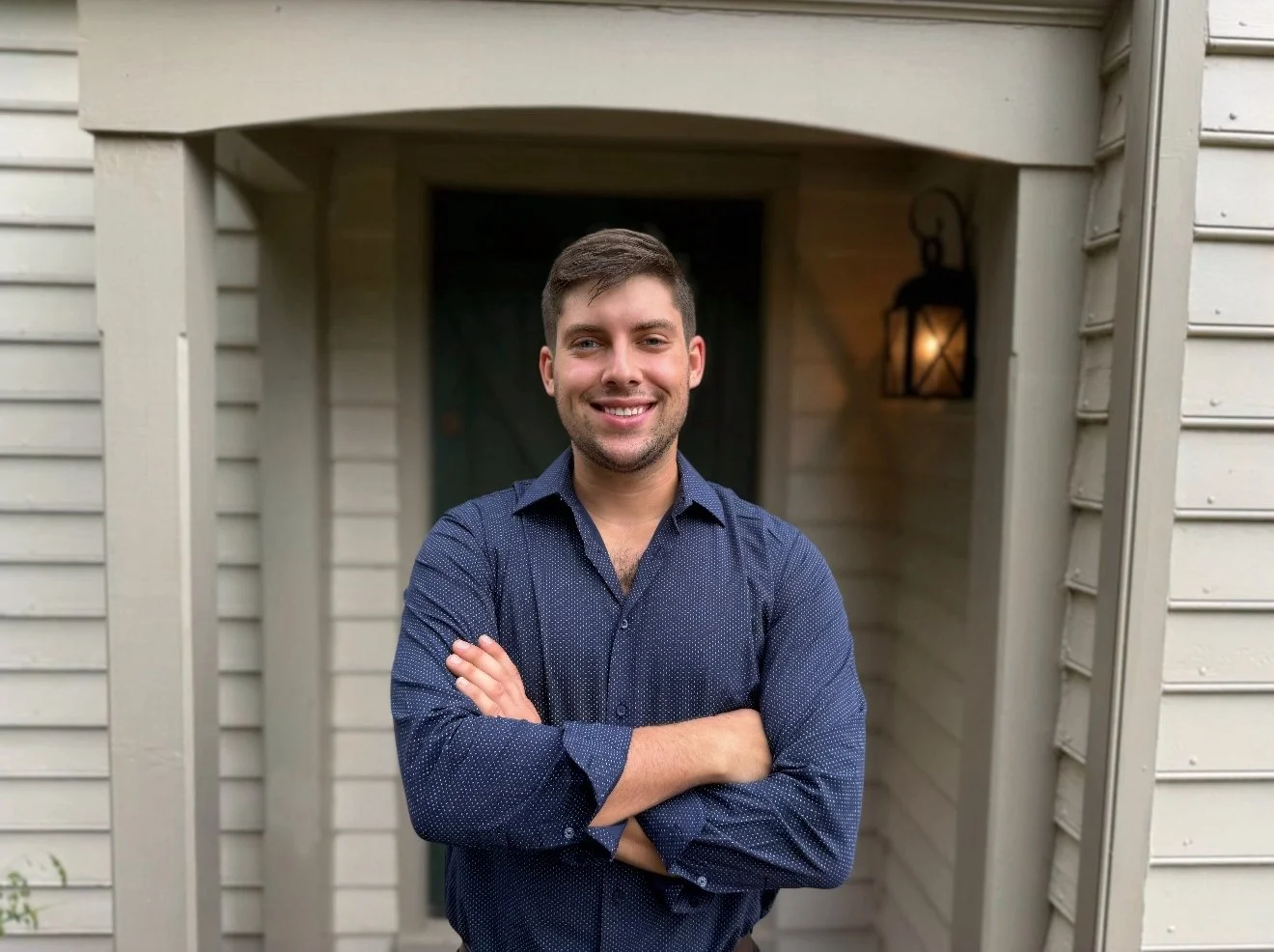 A young man with brown hair smiling and crossing his arms, standing in front of a house with a lit lantern on the wall.