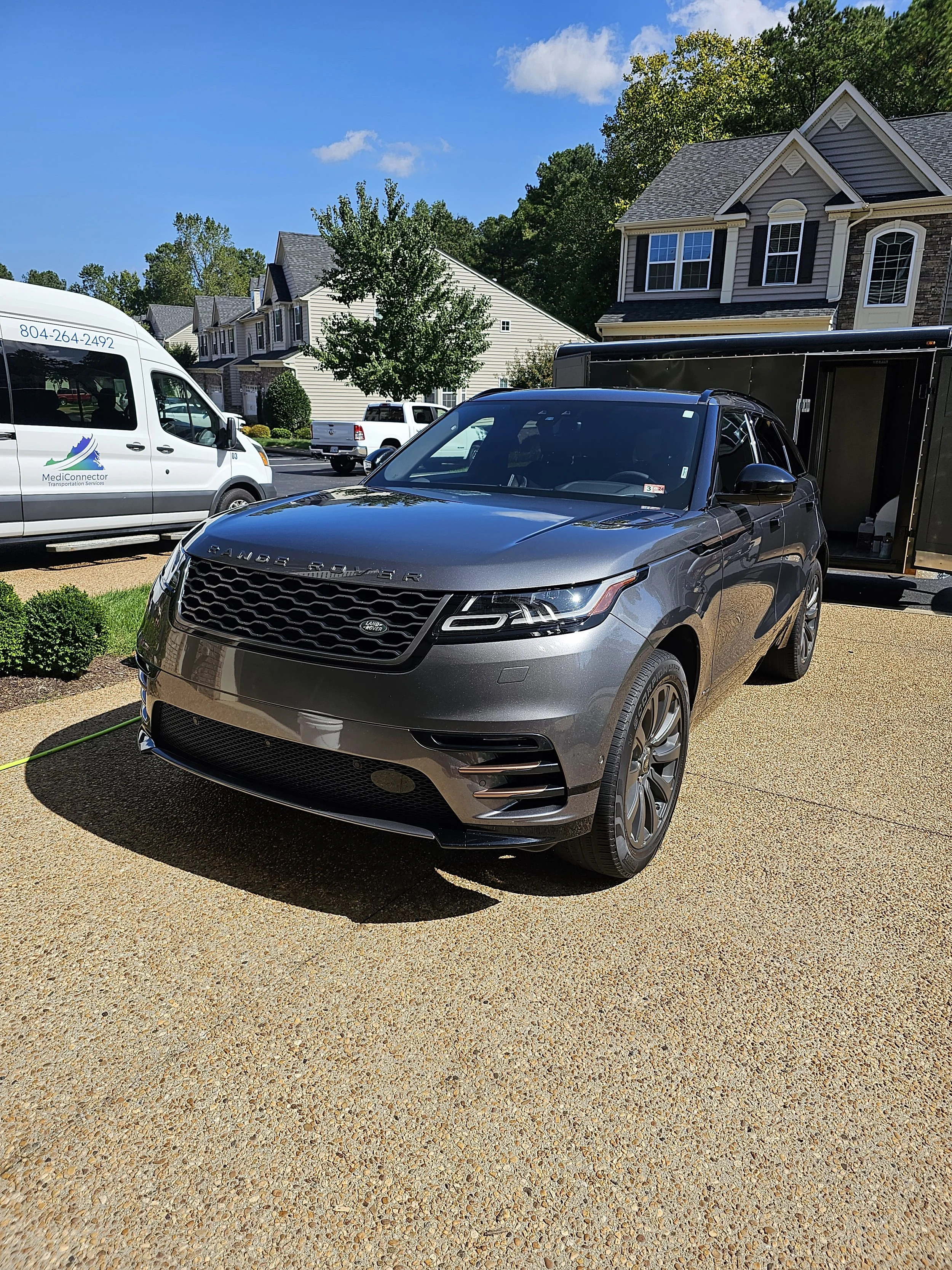 A parking lot with a gray Range Rover, a black trailer, and a white van with MediConnector logo in front of a row of suburban houses with trees on a clear, sunny day.