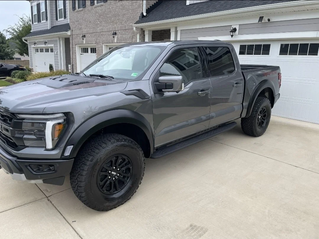 A gray Ford F-150 Raptor pickup truck parked in front of a white garage door on a concrete driveway outside a residential house.