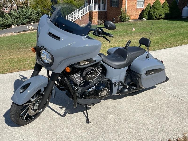 A gray Harley-Davidson touring motorcycle parked on a driveway in front of a grassy yard with bushes and a brick house with stairs.