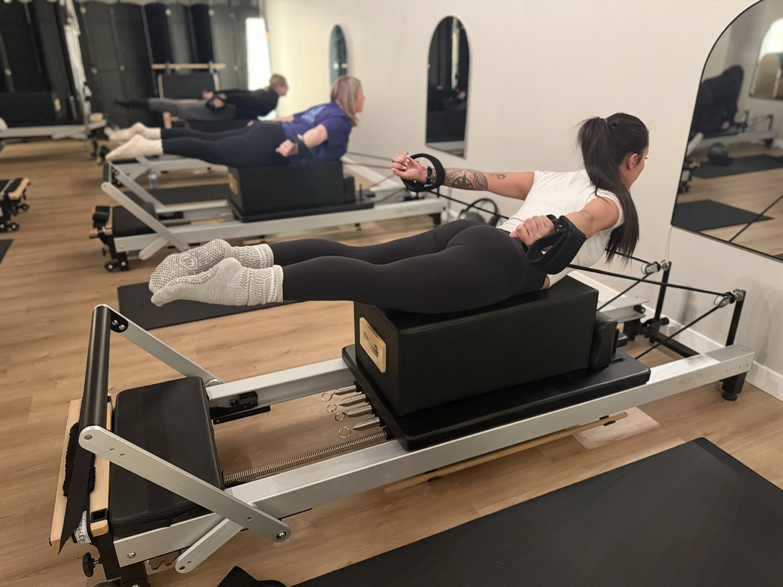 Two women performing back extensions on pilates reformer machines in a fitness studio, with mirrors on the wall.