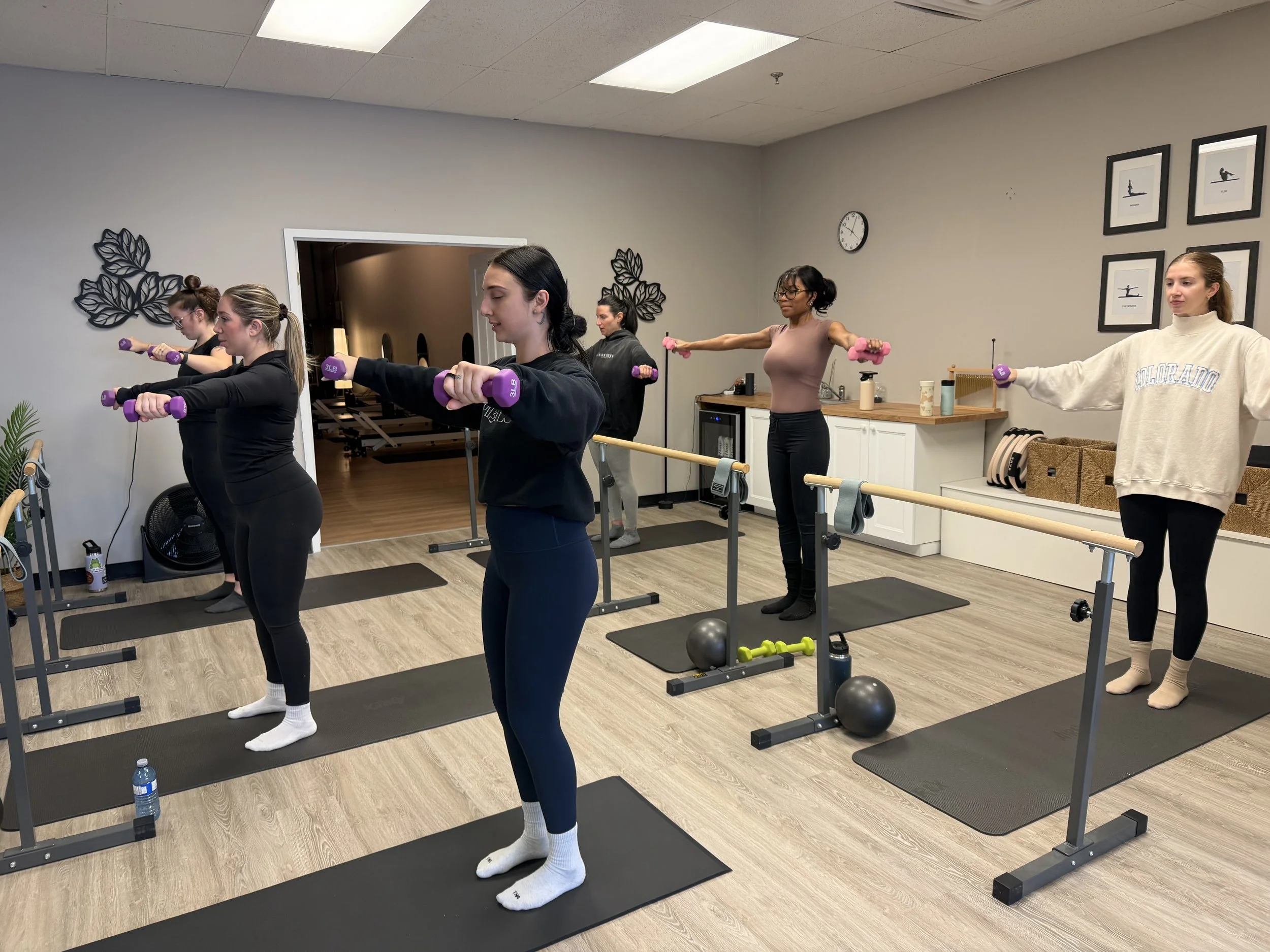 Five women are participating in a fitness class in a gym, holding pink and purple dumbbells out to their sides in a standing exercise. They are positioned on black exercise mats with a wooden ballet barre in front of them. The room has light-colored walls with framed art and a clock, and a doorway leads to another room with workout equipment in the background.