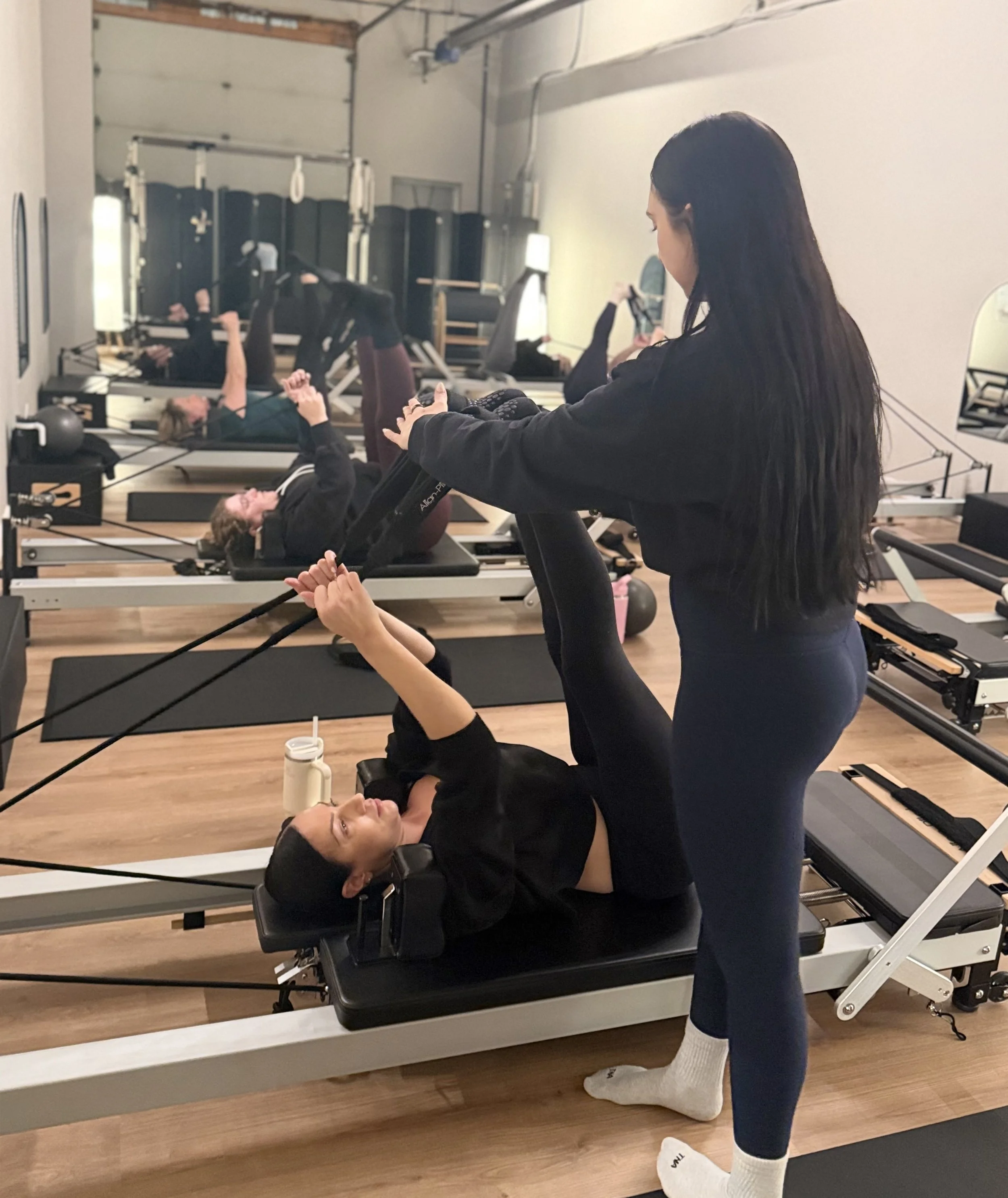 A fitness instructor assists a woman with Pilates exercises on a reformer machine in a gym. In the background, other participants are also working out on reformer machines.