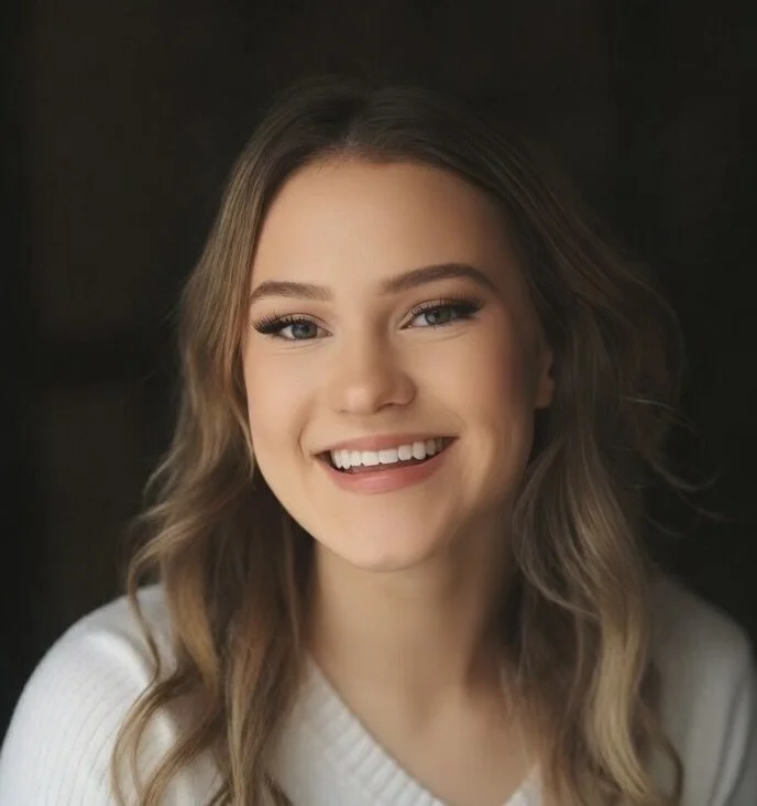 Close-up portrait of a smiling young woman with wavy brown hair, blue eyes, and light makeup, wearing a white top.
