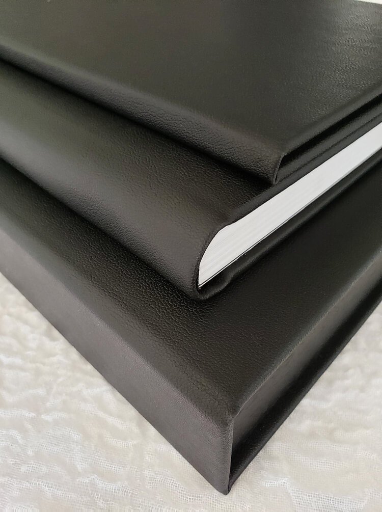 Close-up of a black journal or planner with textured cover, resting on a larger black box or folder, on a white textured surface.
