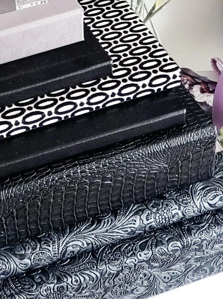 Stacked decorative boxes on patterned surfaces, including a black box with a textured finish and a white box with a black geometric pattern.
