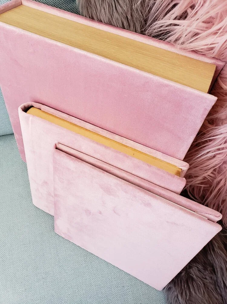 Three pink hardcover books with velvet covers stacked together, leaning against a pink and brown furry background.