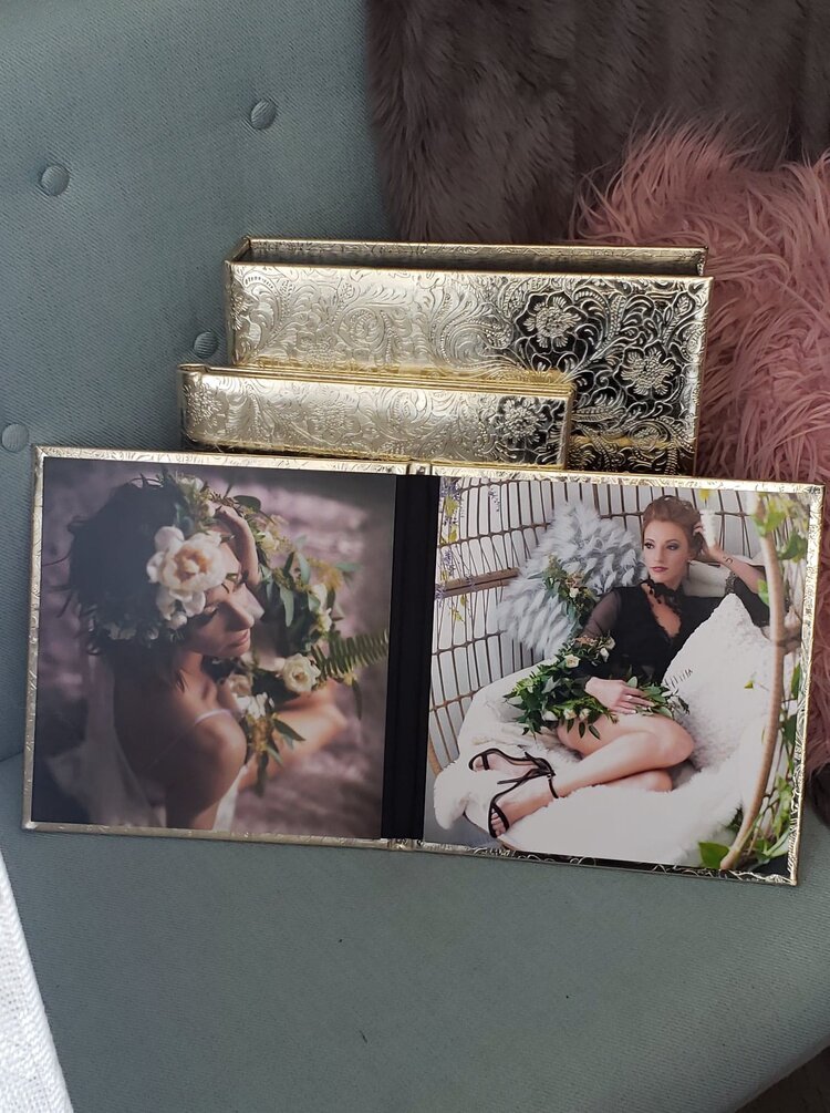 Open photo album with two images of a woman with flowers, one of her wearing a floral crown and the other lounging with flowers, on a grey cushioned surface next to a pink feathered pillow and two decorative gold boxes.
