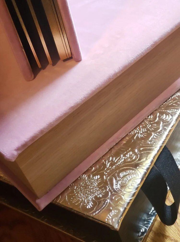 Close-up of a pink book with several other books inside, resting on an ornate gold-colored surface with floral patterns.