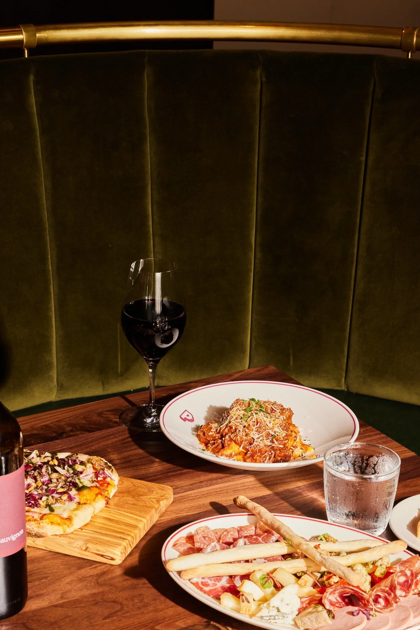 A table with a glass of red wine, a plate of pasta with sauce and grated cheese, a pizza on a wooden board, and a charcuterie platter with various meats, cheeses, and breadsticks, set against a green upholstered bench with a brass railing.