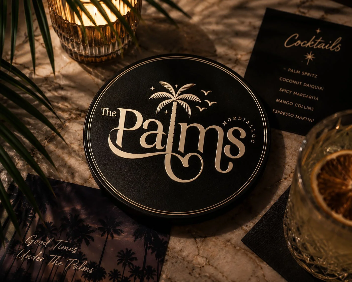 A round black menu with the title 'The Palms' and a palm tree logo, surrounded by glasses with drinks, a black cocktail menu, and greenery, set on a marble surface.