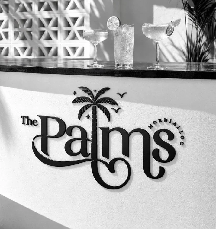 A black and white photo of a bar counter with three cocktails on top, two in margarita glasses and one in a tall glass with a lemon wedge. The bar has a logo that reads "The Palms" with a palm tree illustration, the words "Mordialloc," and some small birds and stars.