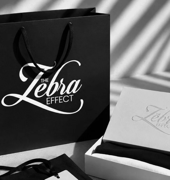 Black shopping bag and white box with the logo for The Lebra Effect, a clothing or accessory brand, on a surface with shadows.