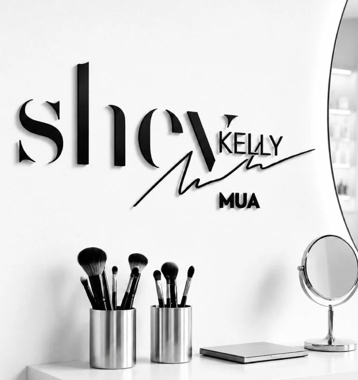 Makeup brushes in silver cups and a mirror on a white vanity against a wall with a sign reading 'slay Kelly MUA'.