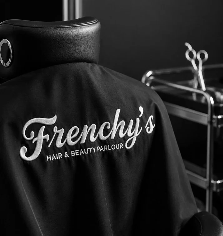 Black cape with 'Frenchy's Hair & Beauty Parlour' embroidered on it hanging in a hair salon, with a styling station and scissors in the background.