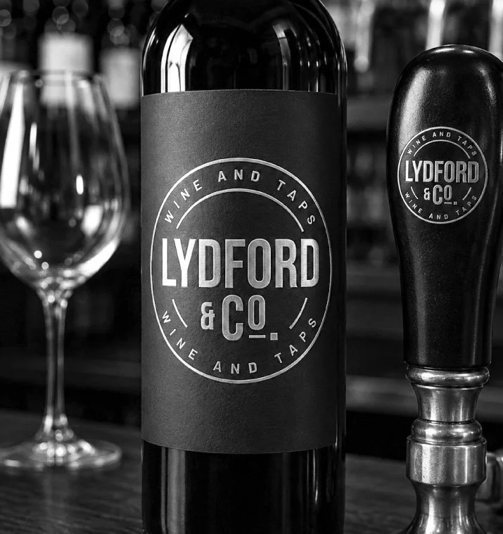 A black wine bottle with a label that reads "Lydford & Co. Wine and Taps," positioned next to a tap handle that matches the bottle's branding, on a wooden surface with a blurred background.