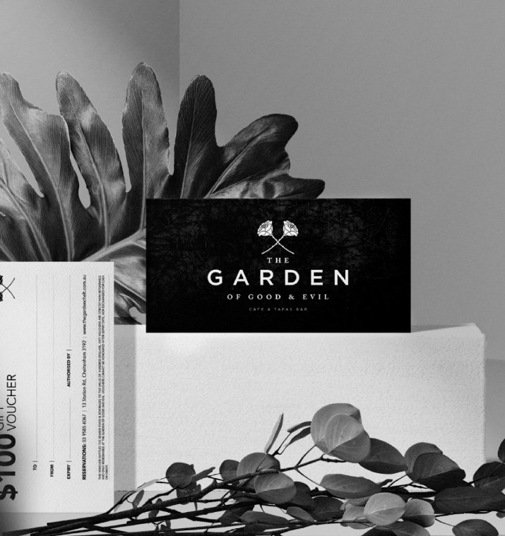 A black and white photo of a sign for The Garden of Good & Evil, a cafe and tapas bar, with large tropical leaves in the background and some foliage in the foreground. A voucher or ticket is partially visible on the left.