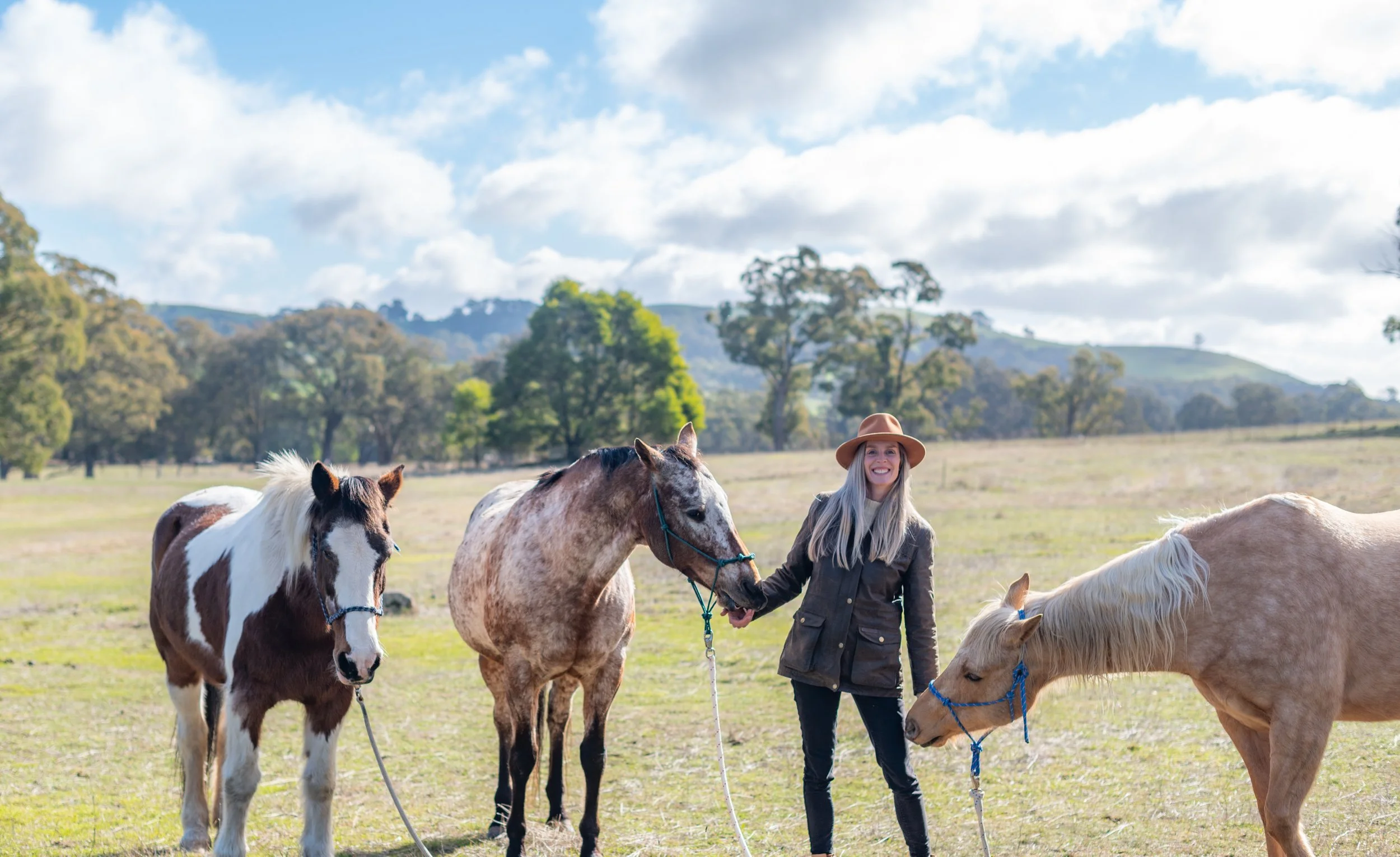 A woman in a brown hat and dark coat smiling while holding the reins of three horses in a grassy field with trees and hills in the background.