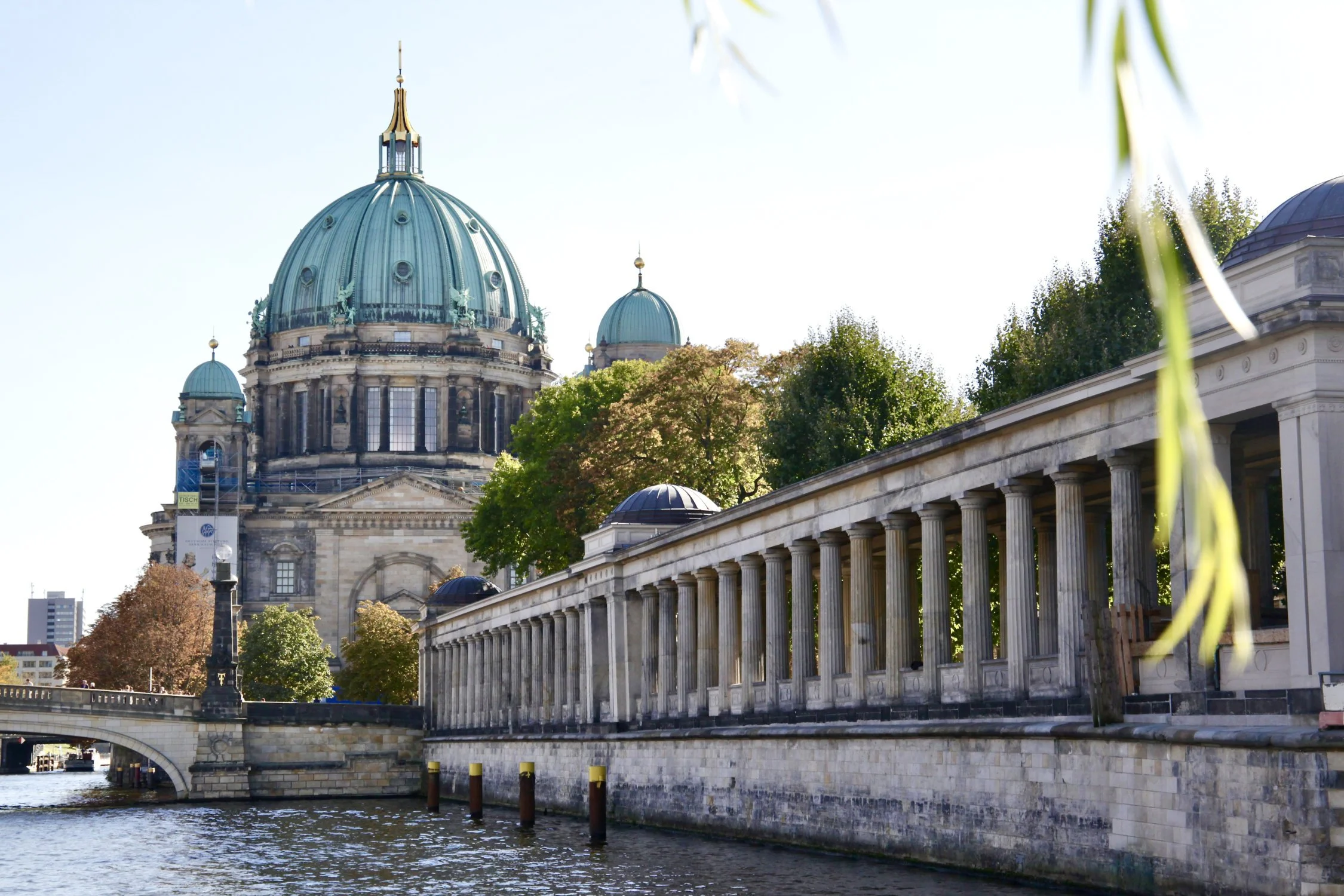 View of the Berliner Dom with green copper domes and gold cross on top. The river Spree is in the foreground and the colonnades on Museum Island are on the right side