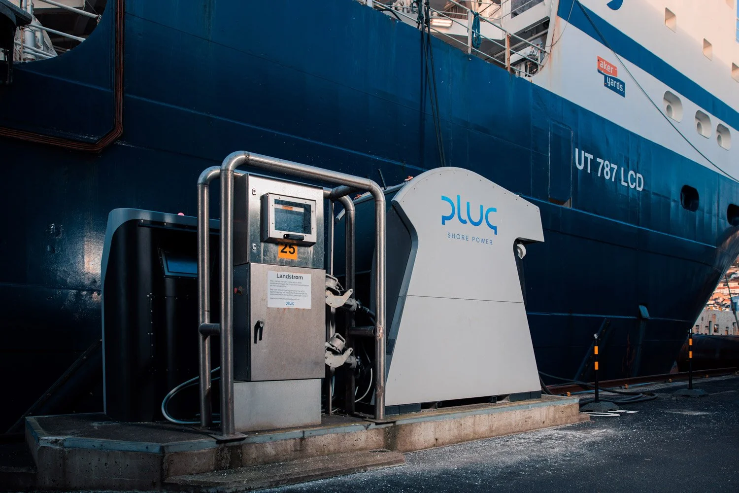 Ladestasjon for elektrisk skip med merke til PLUG Shore Power foran et stort blått cruiseskip.