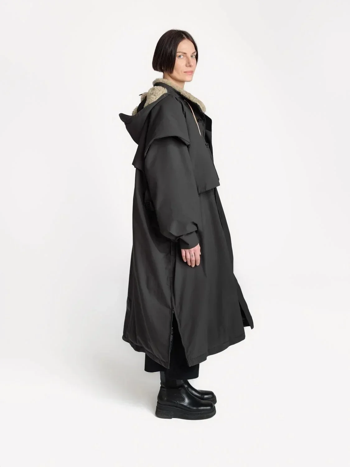 En kvinne med kort mørkt hår i en lang svart opplyst jakke og svarte støvler, står i profil mot en lys bakgrunn.