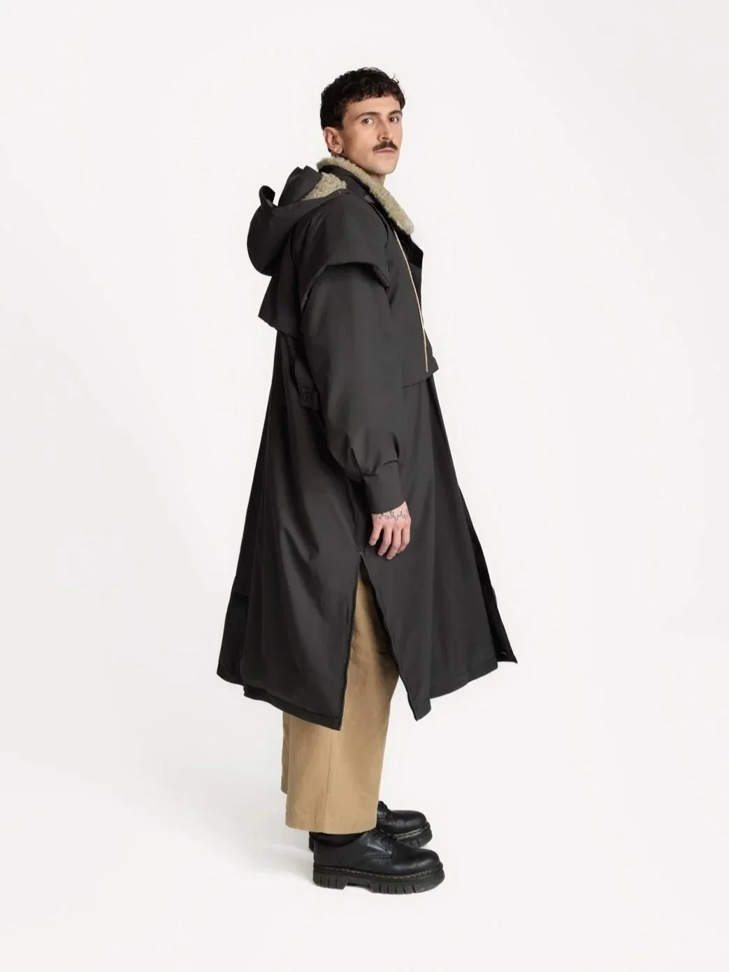 En mann kledd i en lang svart jakke, beige bukser og svarte støvler står mot en hvit bakgrunn.