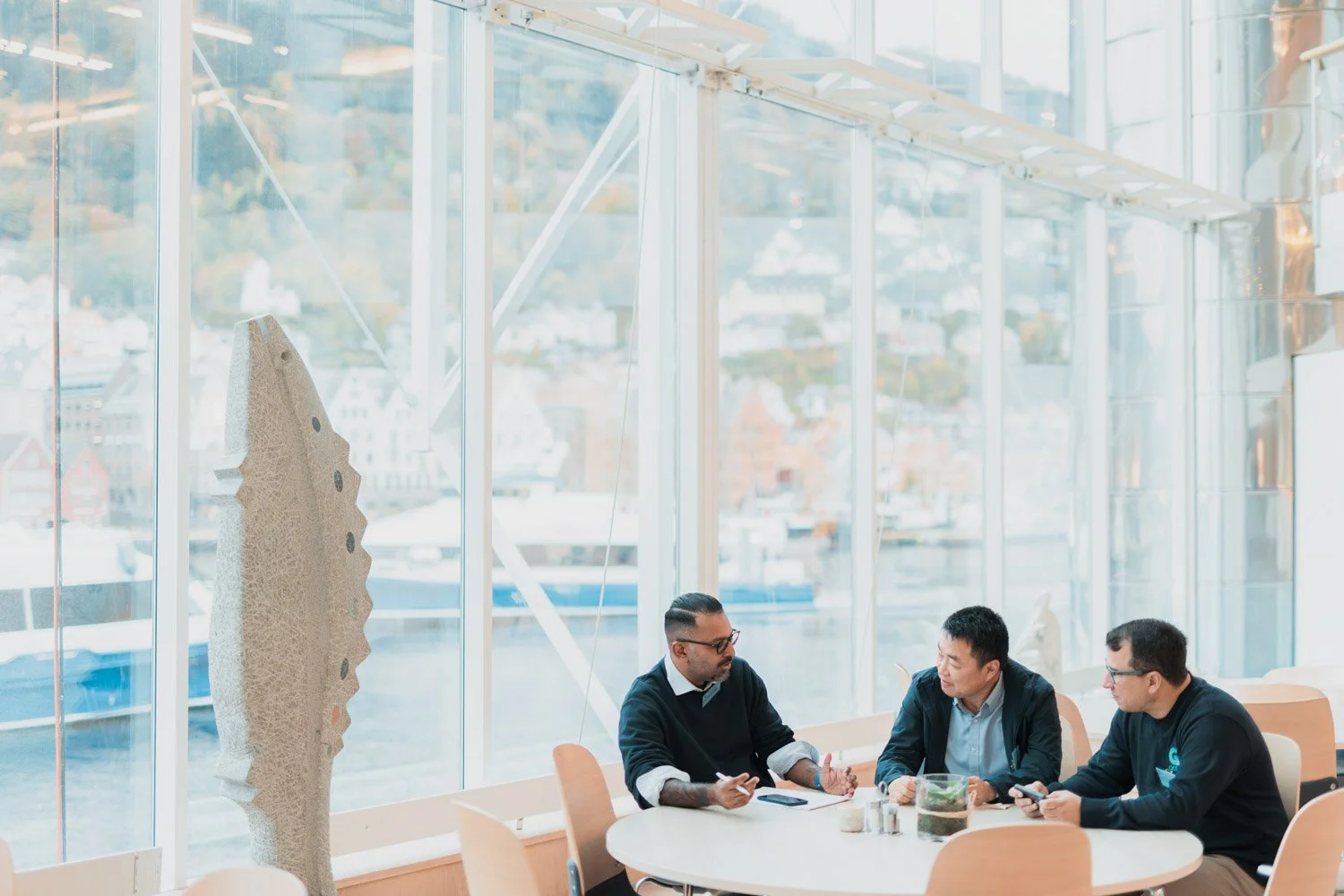 Tre personer som snakker mens de sitter rundt et bord i et moderne rom med store vinduer og utsikt over byen.