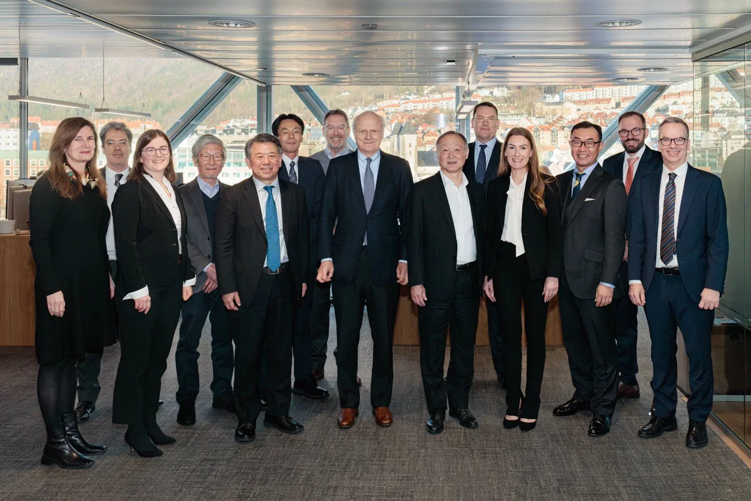 Gruppbilde av menn og kvinner i formelle antrekk i et møterom med stor glassvegg og byutsikt i bakgrunnen.