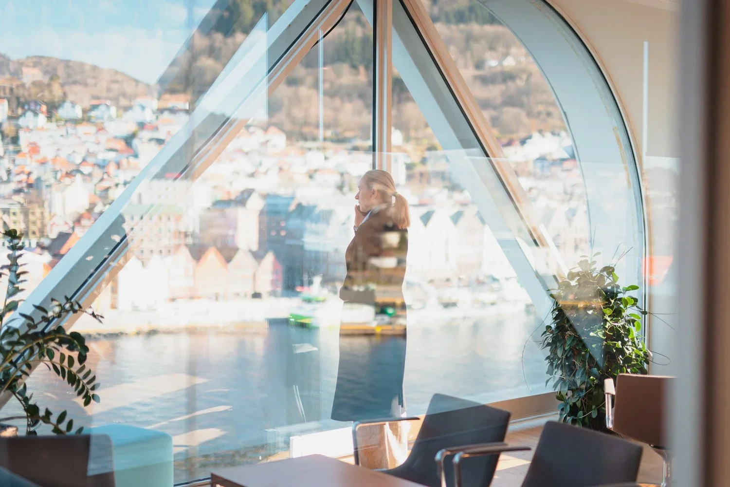 En kvinne står ved et stort glassvindu som vender ut mot en by med fjell i bakgrunnen, og ser ut til å snakke i telefonen. Døren og innendørs planter er også synlige.