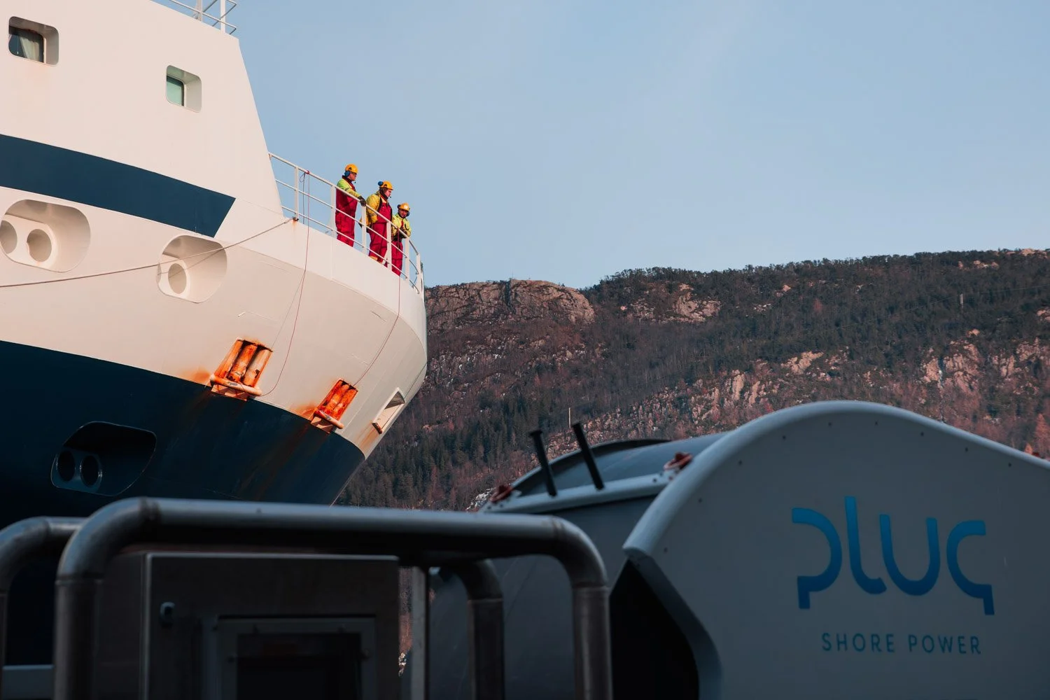 Fire menn i røde og gule redningsvester står på dekk av et stort hvitt cruiseskip ved fjell. Det er en maskin eller utstyr med teksten 'shore power' i forgrunnen, og fjell i bakgrunnen.