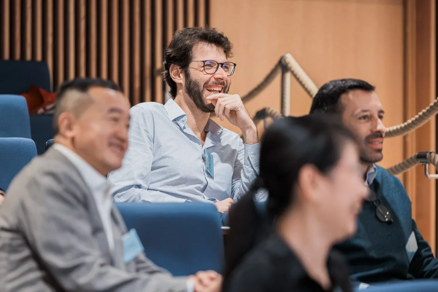 Mann med brunt hår og skjegg ler mens han sitter i en konferansesal med andre deltakere.