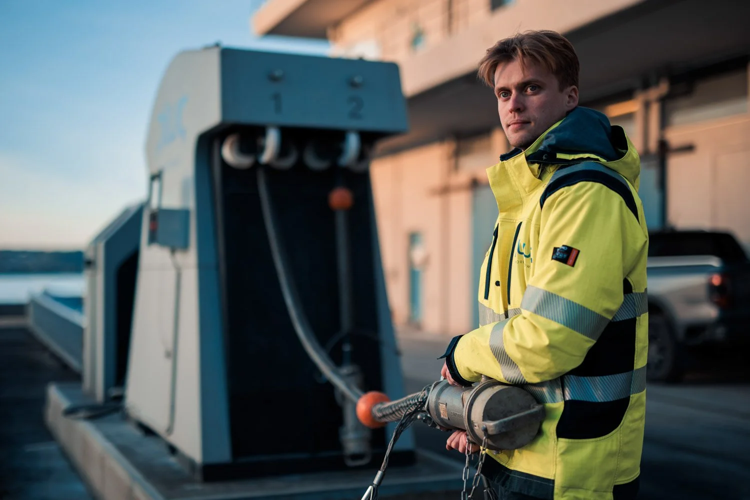 Ung mann i høstjakke med refleksjon holder en elektrisk verktøy, stående ved en datakasse, med en bygning i bakgrunnen.