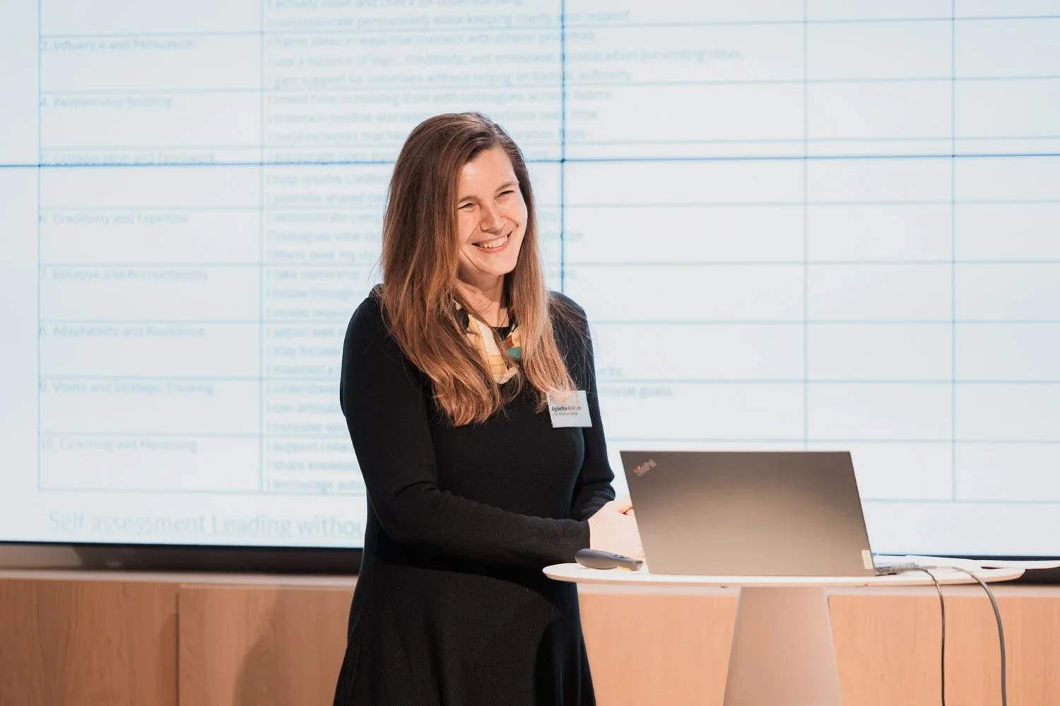 Kvinne med langt brunt hår smiler, står ved en høytaler med åpent bilde av en skjerm med tekst bak seg i et konferanserom.