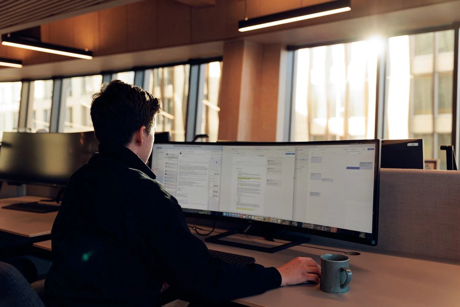 En person som jobber ved en kontorarbeidsplass med doble dataskjermer, med en kaffekopp på pulten, i et rom med store vinduer og naturlig lys.