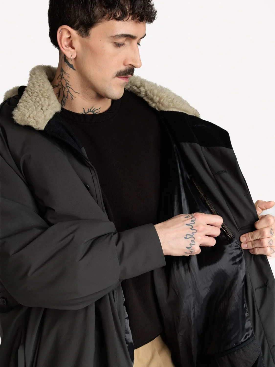 En mann med kort mørkt krøllet hår, skjegg og tatoveringer, iført svart genser og en svart parkas med ullkrage, holder åpen en svart jakke.