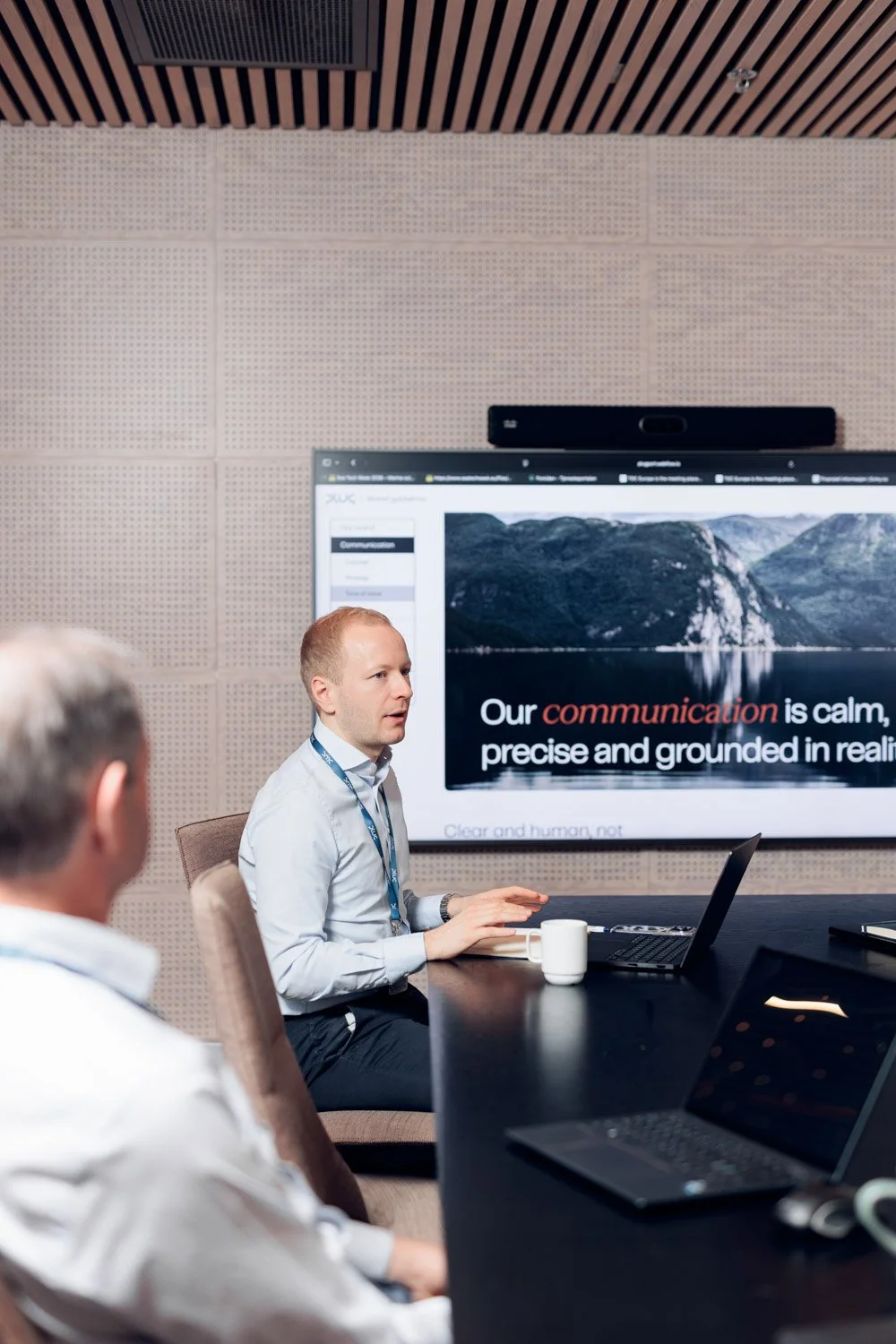 To personer i et møterom med en presentasjon på stor skjerm som viser teksten 'Our communication is calm, precise and grounded in reality'.