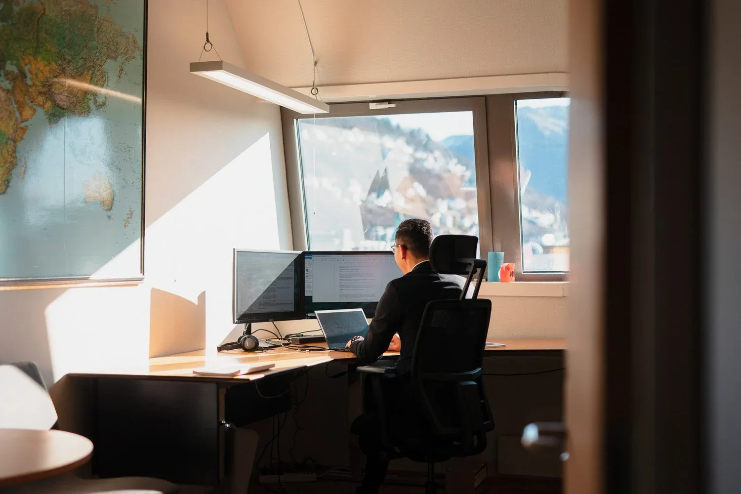 En mann jobber på kontor med to skjermer, en laptop og hodetelefoner. Det er lys fra vinduet bak ham, og et kart av verden på veggen.