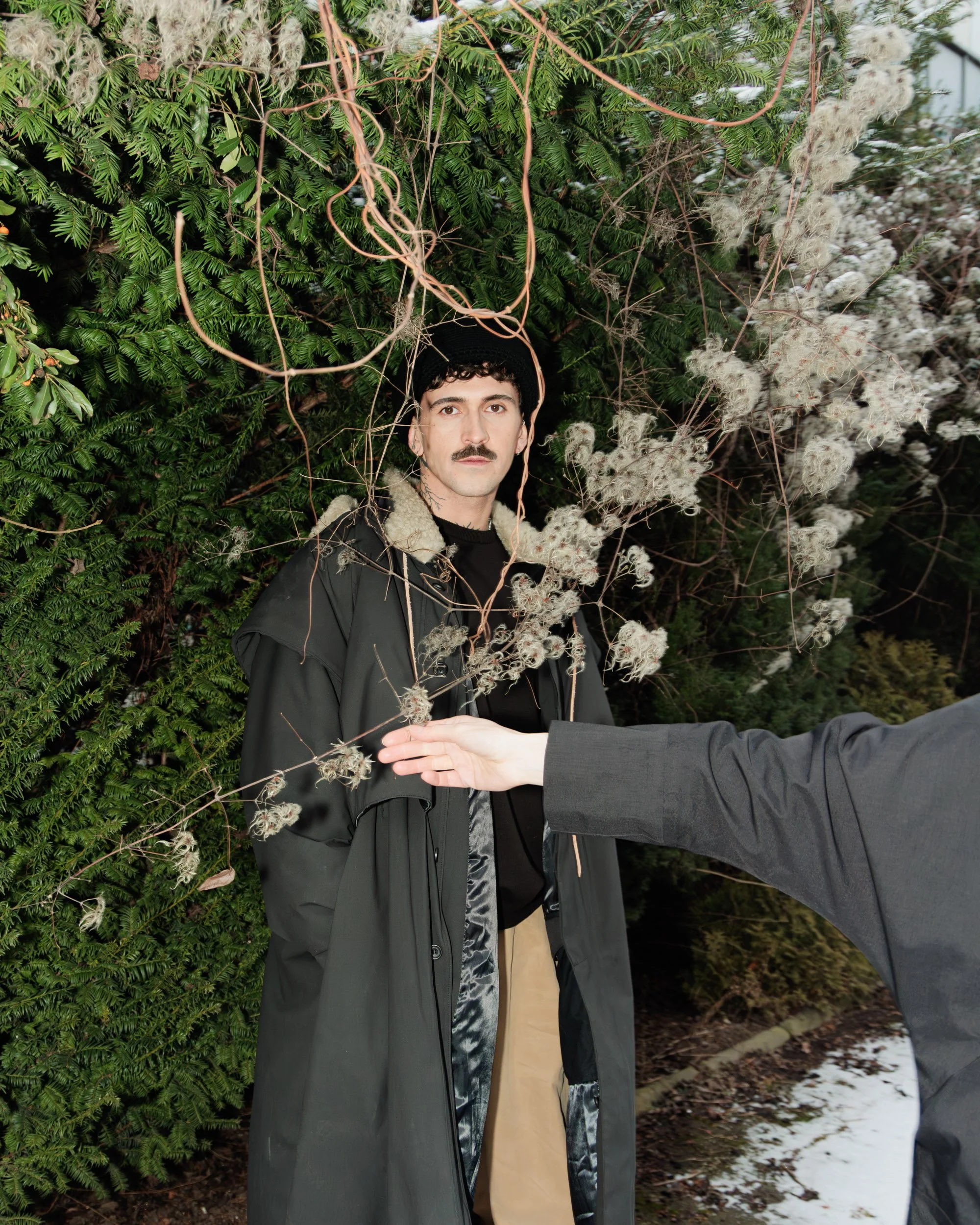 En mann med bart og mørkt krøllet hår står foran en tett busk og ser rett inn i kamera. En person i en grå dress når ut med hånden mot ham.