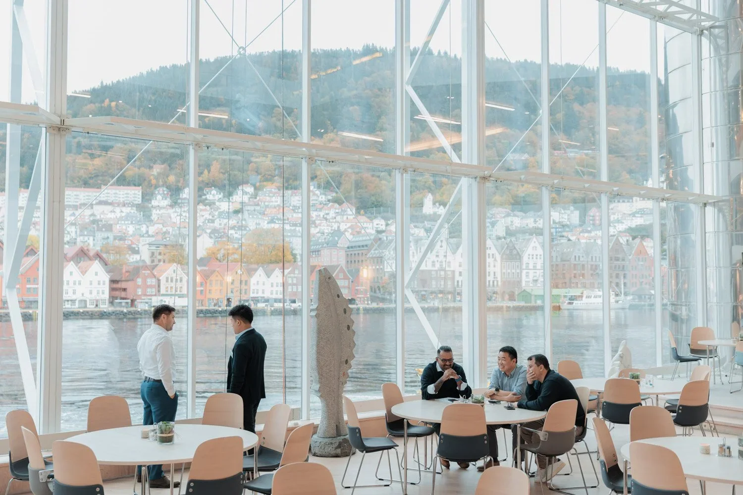 Innendørsområde med store glassvinduer som viser en by og fjell i bakgrunnen, med personer som prater og sitter rundt bord.