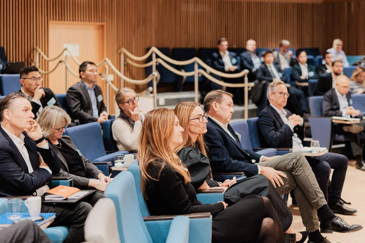 Mennesker i dress på en konferanse eller seminar, sittende i blå stoler i et rom med trepanel på veggen.