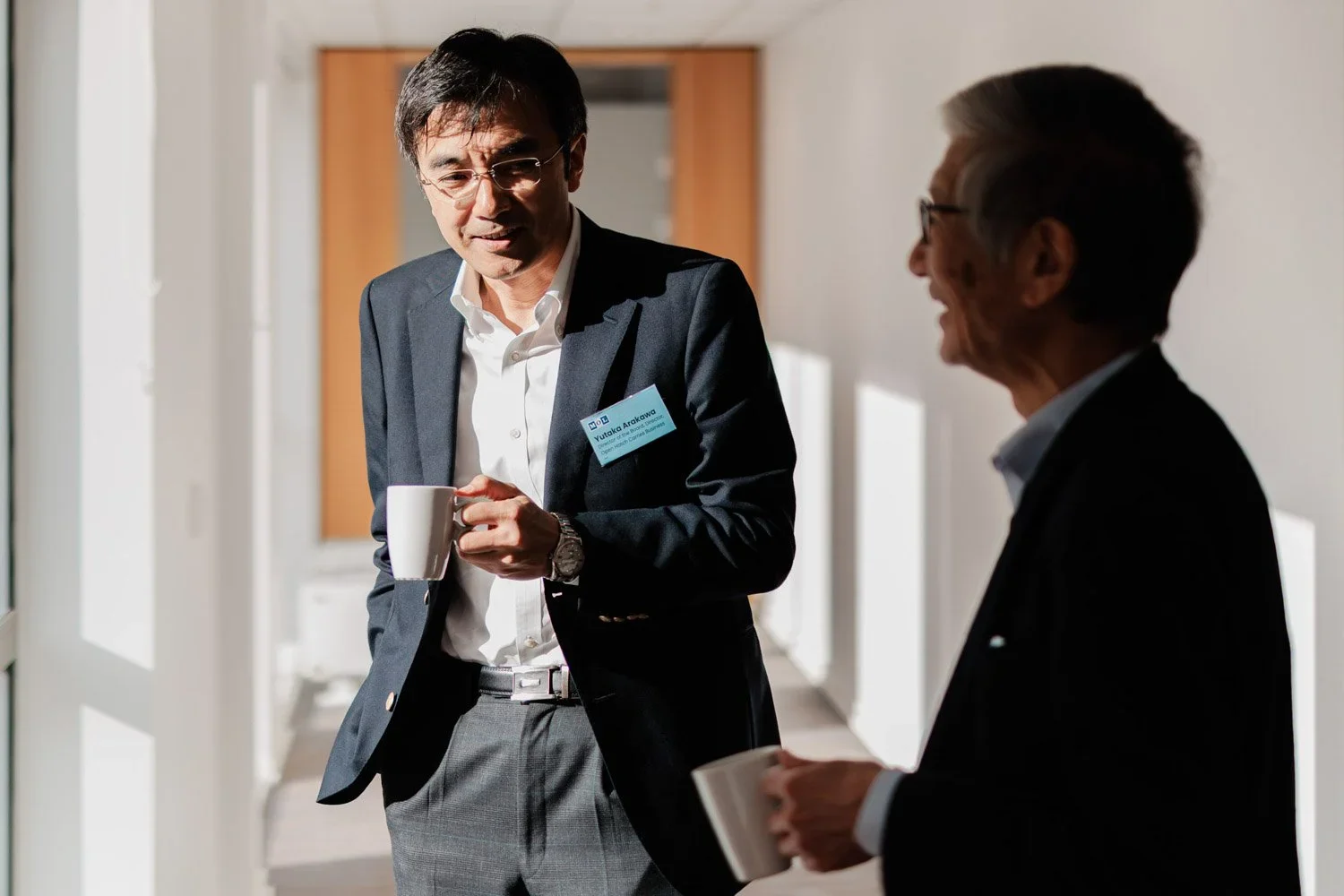 To menn i formelle klær snakker og drikker kaffe i et lyst rom.