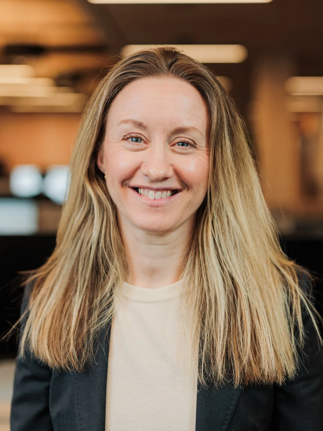 Smilende kvinne med langt blondt hår, iført svart blazer og lysfarget topp, i innendørs miljø.