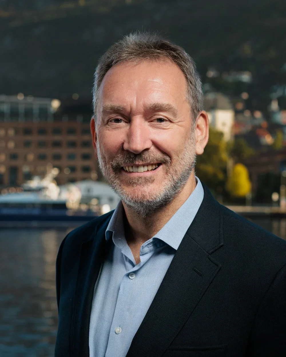 Portrett av en mann med grått hår og skjegg, iført dress og lett blå skjorte, mot en bysiland med vann i bakgrunnen.