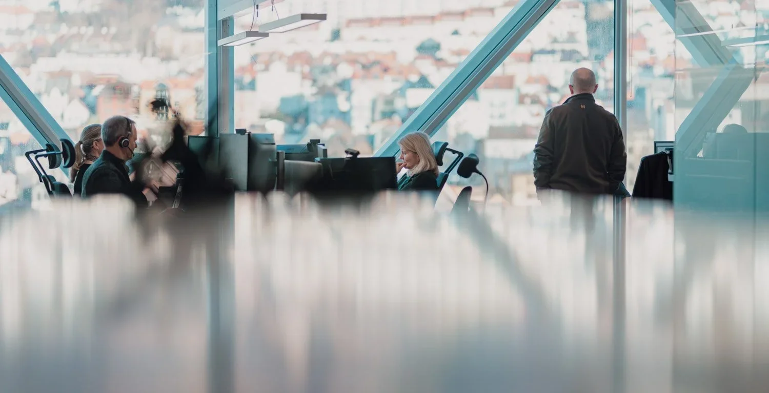 Kontorarbeidsplass med personer som bruker datamaskiner, store vinduer med byutsikt, og to personer som står.