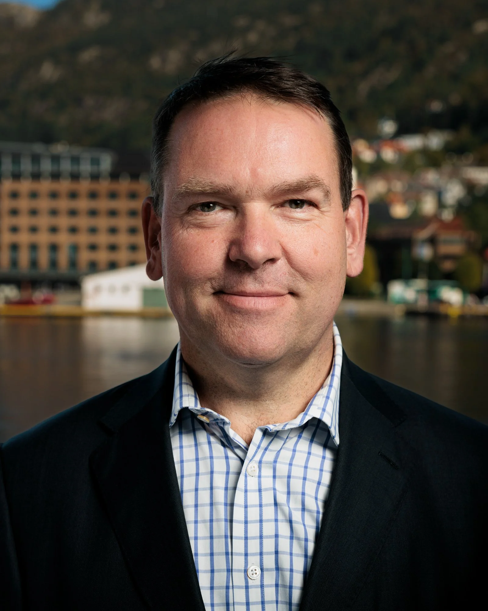 Nærbilde av en mann i dress, stående foran en by ved vannet, med fjell i bakgrunnen.