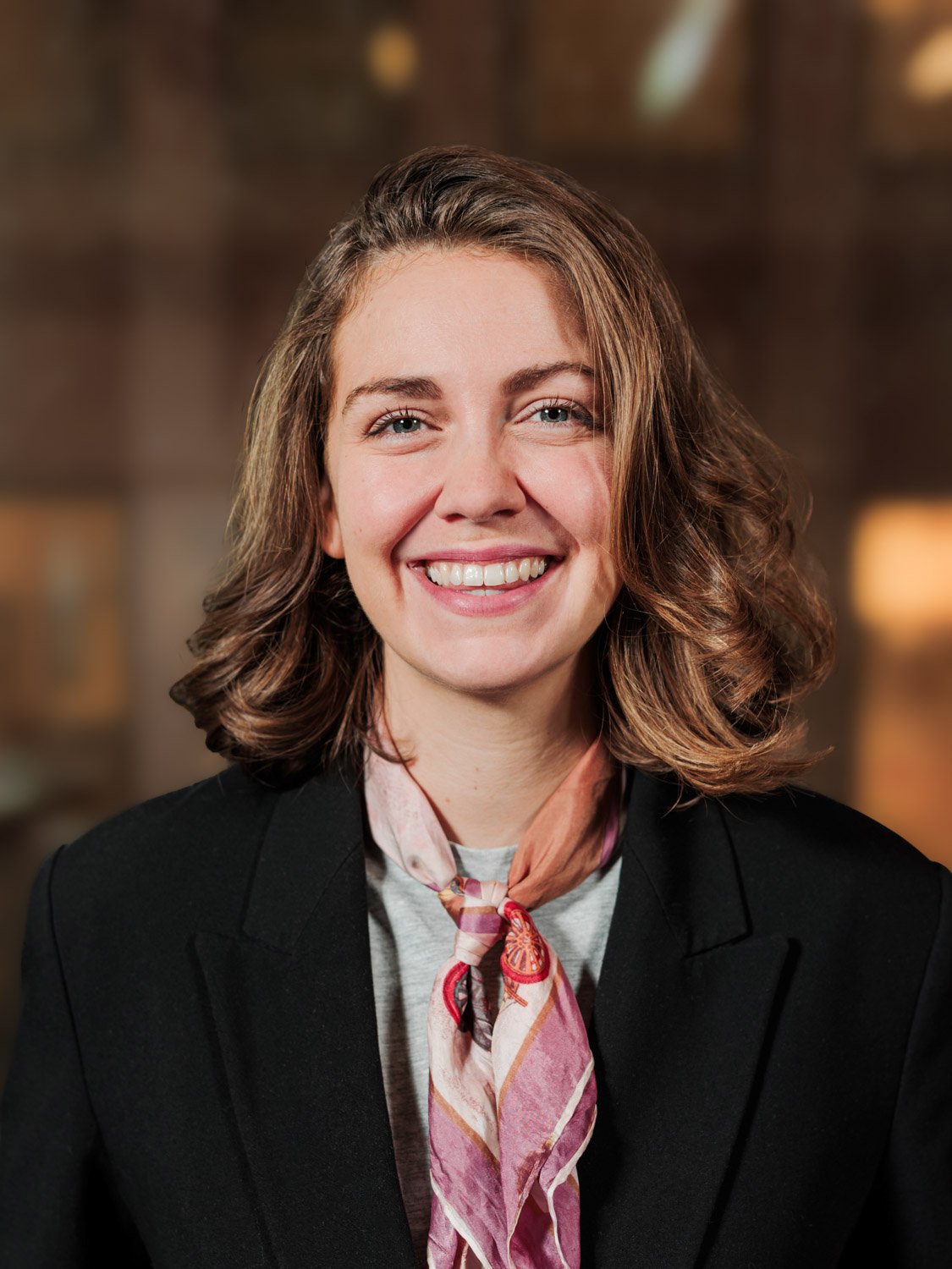 Portrett av en smilende kvinne med brunt, bølget hår, iført svart blazer og rosa skjerf, mot en uklar bar- eller kjøkkenbakgrunn.