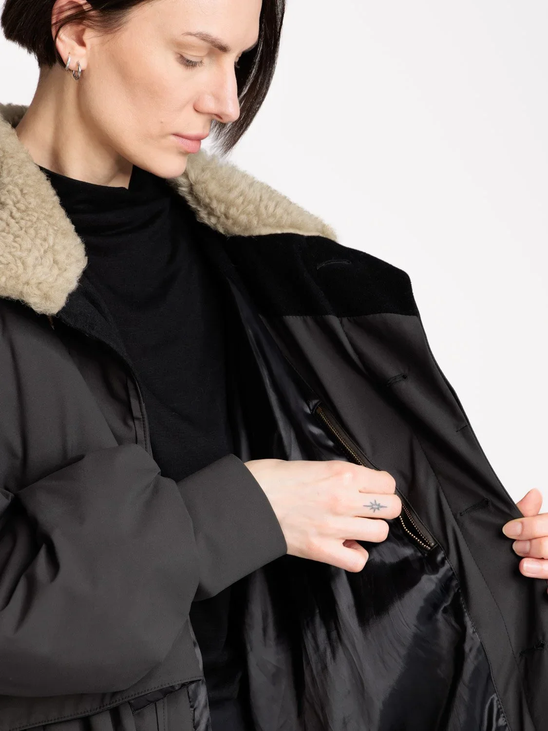 Person som åpner en svart jakke med pelskrage, kledd i svart trøye og har flere ørepynt.